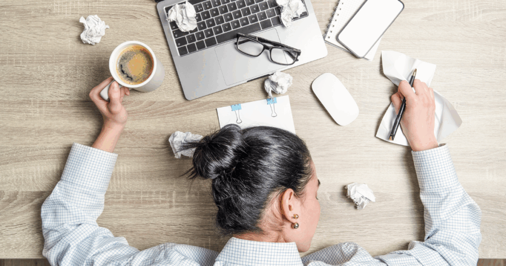 tired woman over 40 laying on her desk with crumbled papers on her desk, cup of coffee, laptop and eyeglasses