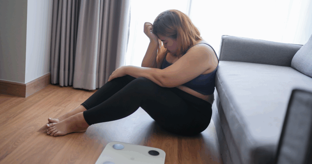overweight asian woman in black workout outfit sitting on floor next to scale in disaapoinment